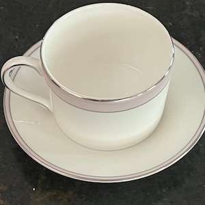 May include: A white teacup and saucer with a silver and pink trim. The cup has a rounded shape and a handle. The saucer has a flat base and a slightly raised rim.