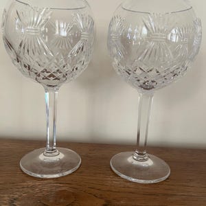 Op de afbeelding: Twee heldere kristallen wijnglazen met ingewikkelde geslepen patronen. De glazen hebben een afgeronde komvorm, een slanke steel en een cirkelvormige voet. Het geslepen glasontwerp heeft een herhalend patroon van stralende lijnen en ruitvormen.