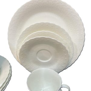 May include: A white porcelain dinner service with a detailed embossed pattern. The set includes a teacup, saucer, and plates of different sizes. The edges of the plates and saucers have a scalloped design.