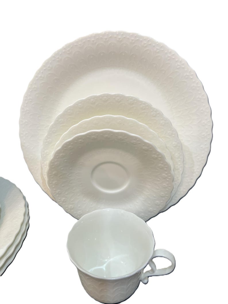 May include: A white porcelain dinner service with a detailed embossed pattern. The set includes a teacup, saucer, and plates of different sizes. The edges of the plates and saucers have a scalloped design.