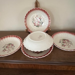 Puede incluir: Conjunto de cinco cuencos de cerámica blanca con borde festoneado y un patrón floral en tonos rosa y rojo. Los cuencos están dispuestos en forma de pirámide sobre una superficie de madera.