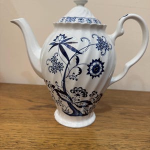 Puede incluir: Tetera de cerámica blanca con un diseño floral azul. La tetera tiene un pico curvo, un asa de bucle y una tapa con un adorno. El patrón floral incluye flores y hojas estilizadas. La tetera está sobre una superficie de madera.