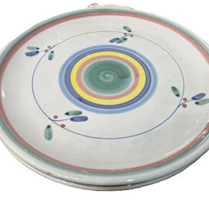 May include: A stack of white ceramic plates with colorful concentric circles in the center. The circles are green, yellow, blue, and pink. The plates have a red and green border and are decorated with blue and green floral accents.