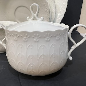 May include: White porcelain sugar bowl with a lid and two handles. The bowl features a decorative embossed pattern of stylized leaves and a scalloped rim. A matching cup and saucer are visible in the background.