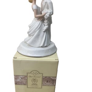 May include: A white porcelain wedding cake topper featuring a bride and groom embracing. The bride wears a flowing gown and the groom a suit. The topper is on a decorative base and sits atop a Lenox box.