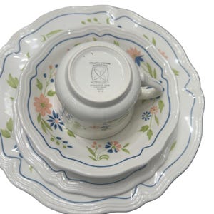 May include: White ceramic dinnerware set with a floral pattern. The set includes a cup, a small plate, a medium plate, and a large plate. The plates have a scalloped edge with a blue trim and floral accents in pink, blue, and green.