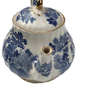 Puede incluir: Una tetera de cerámica blanca con un diseño azul y blanco de patrón Willow. La tetera tiene una boquilla y un adorno de tapa dorados. El asa es azul y blanca, y la tapa tiene un borde dorado.