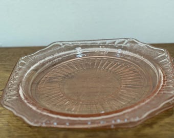 Puede incluir: Un plato de cristal rosa vintage con un borde decorativo festoneado. El plato tiene un centro circular texturizado con líneas radiales. El plato está sobre una superficie de madera.
