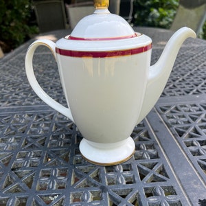 May include: White ceramic coffee pot with a gold trim and a burgundy band around the top. The pot has a gold knob on the lid.