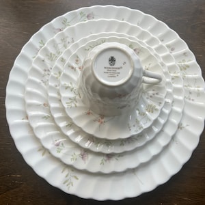 May include: A set of five white ceramic plates with a delicate floral pattern. The plates are nested inside each other, with the smallest plate at the top. The smallest plate has a scalloped edge and features a floral design with pink and green accents. The plates are made by Wedgwood, a well-known British pottery company.