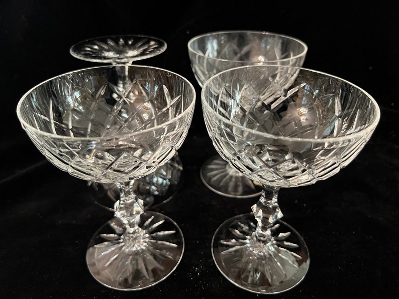 May include: Four clear glass champagne coupes with a cut glass design. The coupes are arranged in a square formation.