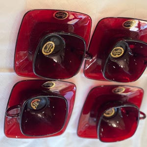 May include: Four red glass teacups and saucers with gold trim. The saucers have a raised edge and the cups have a rounded shape.