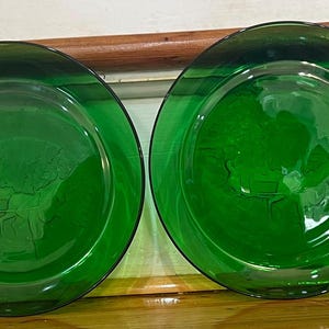 Może przedstawiać: Cztery zielone szklane talerze ze szczegółowym wytłoczonym wzorem. Talerze mają głęboki zielony kolor i klasyczny, elegancki design. Talerze są okrągłe i wydają się być zabytkowe lub antyczne.