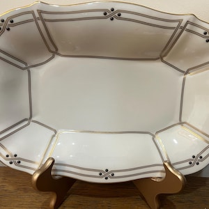 Peut inclure: Un plat de service en porcelaine blanche avec un motif géométrique or et noir. Le plat a un bord festonné et est posé sur un support en bois.