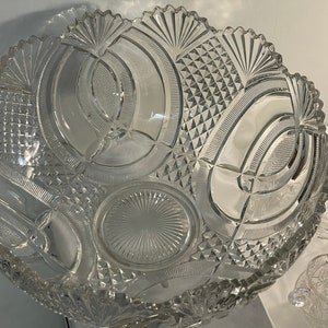 May include: A large, clear glass punch bowl with a cut glass design. The bowl has a scalloped edge and a decorative pattern of circles and arches. The bowl is sitting on a white tablecloth.