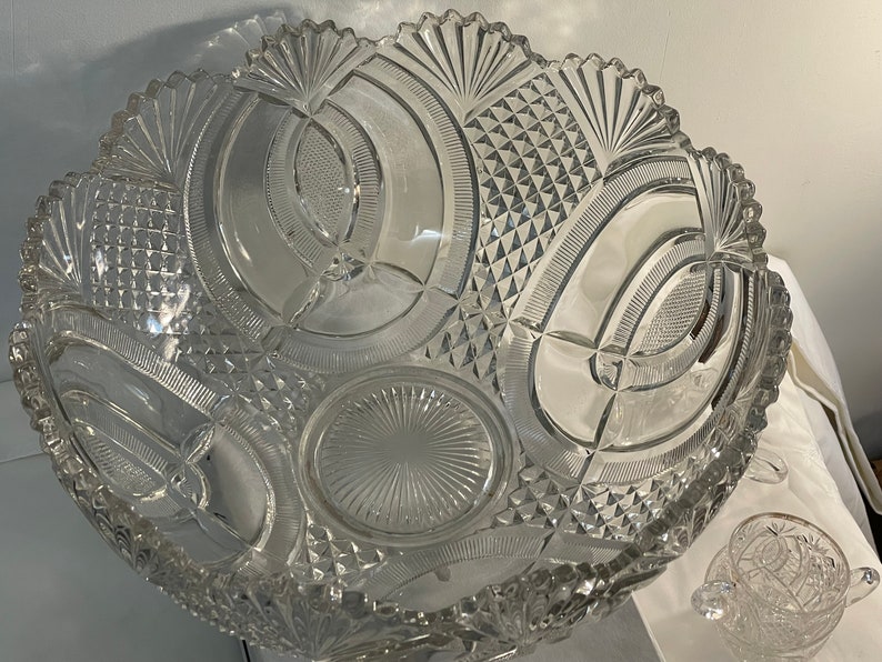 May include: A large, clear glass punch bowl with a cut glass design. The bowl has a scalloped edge and a decorative pattern of circles and arches. The bowl is sitting on a white tablecloth.