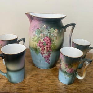 Puede incluir: Una jarra vintage y cuatro tazas con un motivo de uvas. La jarra y las tazas están decoradas con un diseño degradado de rosa, amarillo y azul. Las uvas están pintadas en tonos de rojo y morado.
