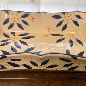 Może przedstawiać: Biała ceramiczna forma do pieczenia z niebieskim i żółtym wzorem kwiatowym. Forma stoi na brązowym metalowym stojaku z uchwytami.