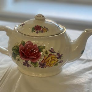 Puede incluir: Una tetera de cerámica blanca con un diseño floral con rosas rojas, amarillas y moradas. La tetera tiene un borde dorado alrededor de la tapa y el pico.