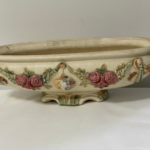 Könnte beinhalten: Cremefarbener Keramik-Blumentopf mit ovaler Form und floralem Relief. Mit roten Rosen, grünen Blättern und einem Cherub-Design. Er hat verzierte Griffe und einen Sockel. Der Topf ist ca. 40 cm lang.