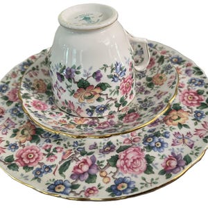 May include: A white porcelain teacup, saucer, and plate set. The set features a floral pattern with pink, purple, blue, and yellow flowers. The edges of the plates and saucer are trimmed with gold. The teacup is upside down on the saucer.