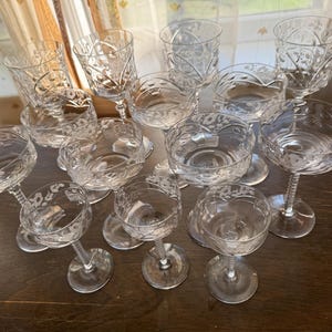 Puede incluir: Una colección de 13 vasos de cristal transparente con tallo y diseños florales grabados. Los vasos están dispuestos sobre una superficie de madera.