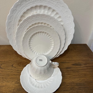 Puede incluir: Un juego de cuatro platos de cena de cerámica blanca con un delicado patrón de encaje. Los platos están apilados uno encima del otro, con una taza de té y un platillo blancos a juego en la parte superior. La taza de té tiene la palabra "Spode" impresa en la parte inferior.
