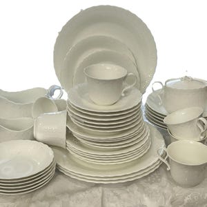 May include: A full white porcelain dinner service. The set includes plates, bowls, cups, saucers, a sugar bowl, and serving jugs. The dishes have a textured, embossed design. The set is arranged on a white tablecloth.