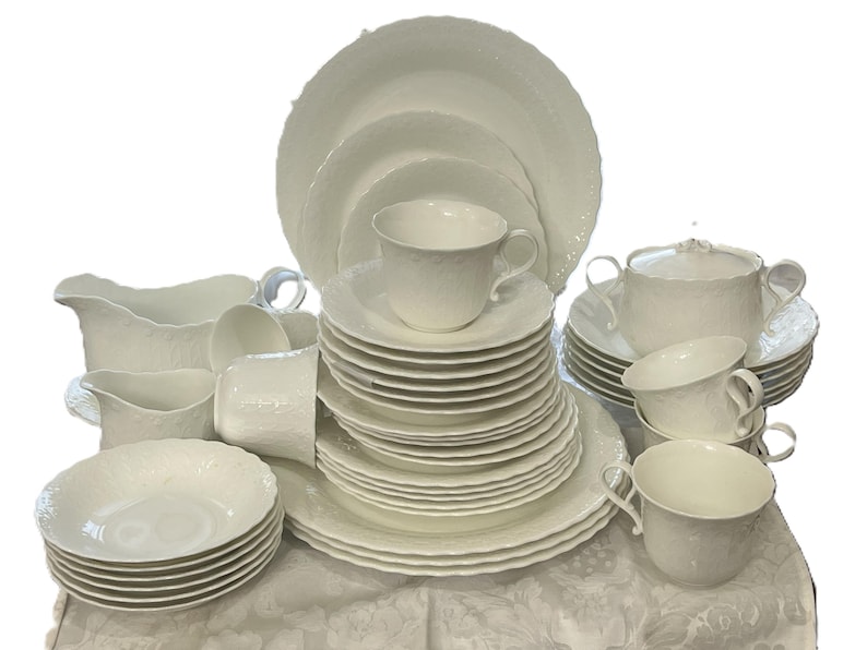May include: A full white porcelain dinner service. The set includes plates, bowls, cups, saucers, a sugar bowl, and serving jugs. The dishes have a textured, embossed design. The set is arranged on a white tablecloth.