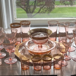 May include: A collection of pink and gold glassware, including wine glasses, champagne flutes, and small bowls. The glassware is arranged on a table with a gold-trimmed tablecloth.