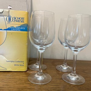 K&ouml;nnte beinhalten: Vier klare Weingl&auml;ser aus Glas, von denen drei leer sind und eines mit Wei&szlig;wein gef&uuml;llt ist. Die Gl&auml;ser stehen auf einer Holzoberfl&auml;che. Der Karton im Hintergrund ist mit "Schott Zwiesel The Connoisseur's Crystal Lexington Collection White Wine 4" beschriftet.
