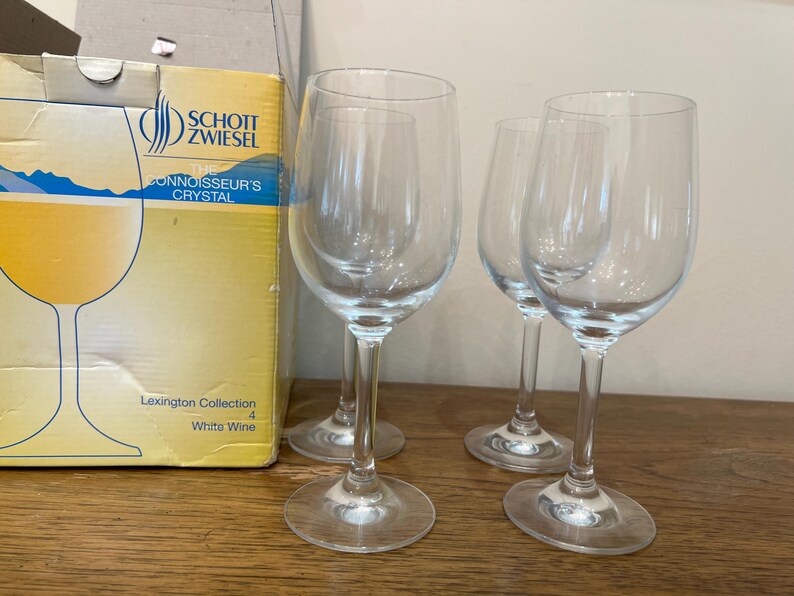 K&ouml;nnte beinhalten: Vier klare Weingl&auml;ser aus Glas, von denen drei leer sind und eines mit Wei&szlig;wein gef&uuml;llt ist. Die Gl&auml;ser stehen auf einer Holzoberfl&auml;che. Der Karton im Hintergrund ist mit "Schott Zwiesel The Connoisseur's Crystal Lexington Collection White Wine 4" beschriftet.