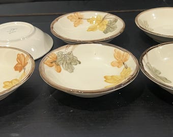 Pode incluir: Um conjunto de cinco tigelas redondas de cerâmica com uma base de cor creme e uma borda castanha. Cada tigela apresenta um padrão pintado de folhas de outono em tons de laranja, amarelo e verde suave. Uma tigela está virada para baixo.