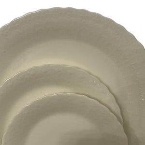 May include: Three off-white ceramic plates with scalloped edges are stacked. The plates have a textured, embossed pattern. The plates are nested, with the largest plate at the bottom and the smallest at the top. The plates are clean and simple.