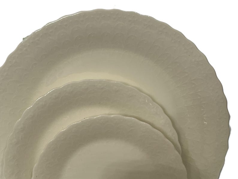 May include: Three off-white ceramic plates with scalloped edges are stacked. The plates have a textured, embossed pattern. The plates are nested, with the largest plate at the bottom and the smallest at the top. The plates are clean and simple.