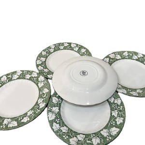 Może przedstawiać: Zestaw białych owalnych talerzy z zielono-białym wzorem liści na obrzeżach. Jeden talerz jest odwrócony, ukazując stempel na spodzie. Talerze ułożone na białym tle.