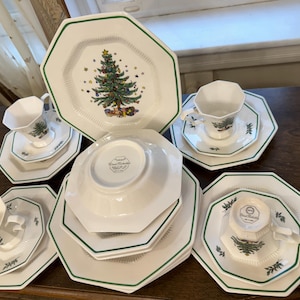 May include: A set of white octagonal Christmas themed dinnerware with green trim. The set includes a large dinner plate, a smaller plate, a bowl, and a teacup and saucer. The dinner plate features a green Christmas tree with red and gold ornaments.