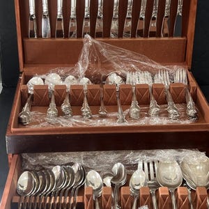 Könnte beinhalten: Ein silberfarbenes Besteckset, angeordnet in einem zweistöckigen Holzkasten mit braunem Samtfutter. Das Set enthält Messer mit verzierten Griffen, verschiedene Löffel und Gabeln, teilweise in durchsichtige Kunststofffolie eingewickelt.