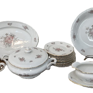 May include: A complete white porcelain dinnerware set with a floral pattern in pink and purple. The set includes serving platters, bowls, plates, a gravy boat, a creamer, and a lidded soup tureen. Gold trim accents the edges.