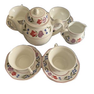 Könnte beinhalten: Ein weißes Keramik-Teeservice mit floralen Designs in Rot, Lila und Blau. Das Set umfasst eine Teekanne, einen Milchgießer, eine Zuckerdose und Tassen mit Untertassen. Die Teekanne hat eine runde Form mit Deckel und Henkel. Die Tassen und Untertassen haben einen gewellten Rand.