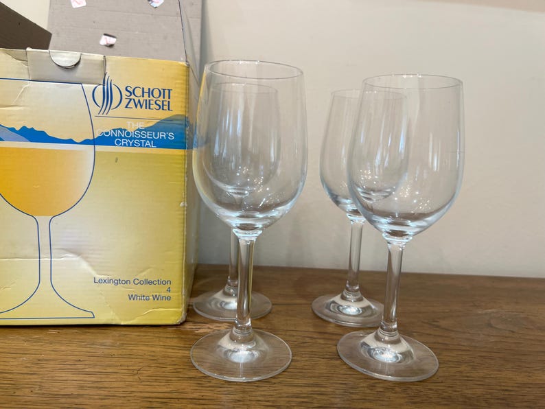 K&ouml;nnte beinhalten: Vier klare Weingl&auml;ser aus Glas f&uuml;r Wei&szlig;wein auf einer Holzoberfl&auml;che. Die Gl&auml;ser stammen aus der Lexington Collection von Schott Zwiesel. Das Logo Connoisseur's Crystal ist auf dem Karton im Hintergrund zu sehen.