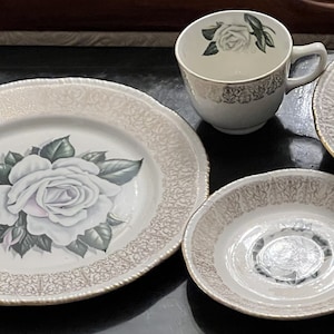 Pode incluir: Um conjunto de louça de porcelana branca com acabamento dourado e um desenho de rosa branca. O conjunto inclui um prato grande, um prato pequeno, uma tigela e uma chávena.