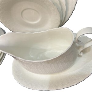 May include: A white porcelain gravy boat with a matching saucer. The boat has a curved handle and a scalloped rim. The set has a textured, embossed pattern. Several matching plates are visible in the background.