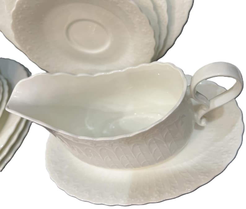 May include: A white porcelain gravy boat with a matching saucer. The boat has a curved handle and a scalloped rim. The set has a textured, embossed pattern. Several matching plates are visible in the background.