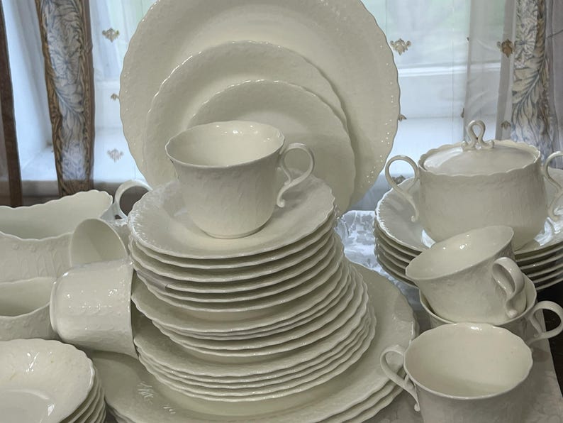 May include: A complete white porcelain dinner service, featuring plates, cups, saucers, and a lidded serving bowl. The items have a textured finish and a decorative scalloped edge. The set is arranged on a table, ready for use.