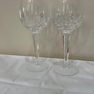 May include: Two clear crystal wine glasses with intricate cut patterns. The glasses have a classic goblet shape with a round base and a long, slender stem. The background is a plain white surface.