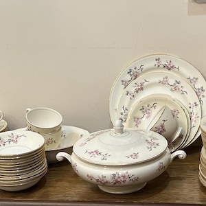 Könnte beinhalten: Ein komplettes Vintage-Porzellan-Geschirrset. Das Set umfasst Teller, Schalen, Tassen, Untertassen, eine Servierplatte, eine Terrine und eine Sauciere. Jedes Stück ist weiß mit einem zarten Blumenmuster in Rosa- und Lilatönen und Goldrand.