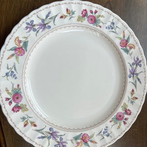 May include: A white ceramic dinner plate with a floral design. The border features a delicate pattern of pink, purple, and yellow flowers with green leaves. The plate has a scalloped edge.