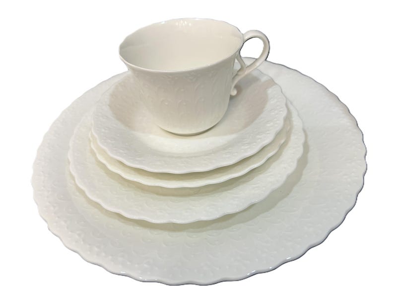 May include: A white porcelain teacup and saucer set with a delicate embossed floral pattern. The set includes a teacup, a saucer, a small bowl, and two plates. The edges of the plates and bowls have a scalloped design.