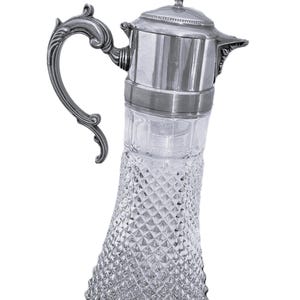 May include: A silver-plated and cut-glass pitcher with a lid and ornate handle. The clear glass body features a diamond-cut pattern. The pitcher is likely used for serving beverages and adds a touch of elegance to any table setting.